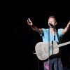 Chris Martin performs on stage with his arms outstretched, his guitar hanging on his neck and singing into a microphone