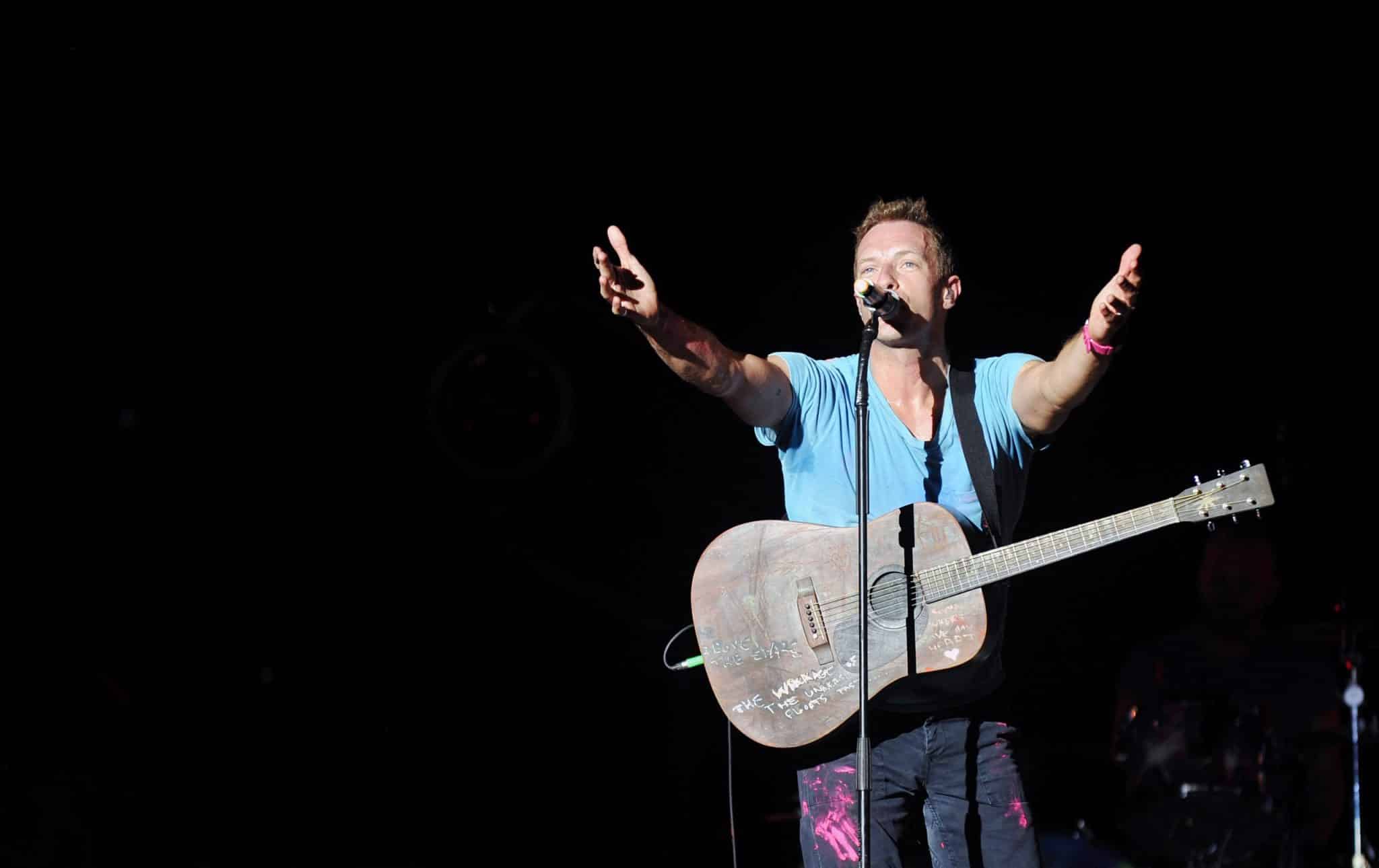 Chris Martin performs on stage with his arms outstretched, his guitar hanging on his neck and singing into a microphone