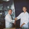 Chef Daniel Boulud with Blake LIvely, both in a kitchen wearing chef's gear and smiling. He is rolling up her sleeve.